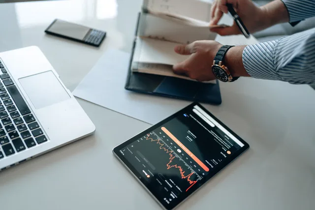Professional workspace featuring a laptop, tablet with analytics, and a person organizing documents. Checking budget approval workflows in marketing software