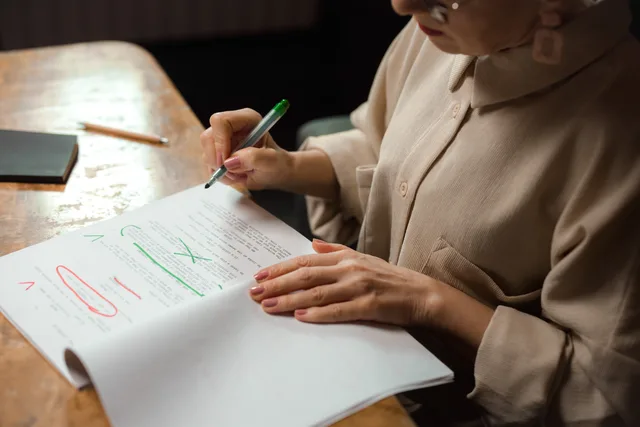 Close-up of a senior woman editing a manuscript at a desk with a green marker indoors. Comparing editorial workflows in CMS products: corrections